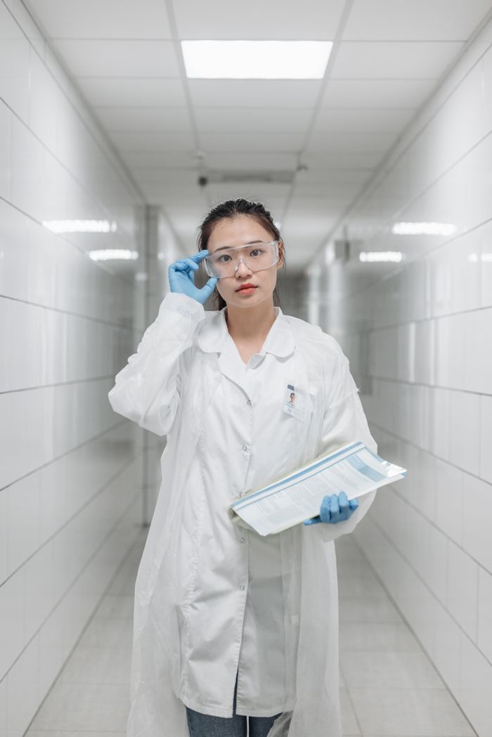 A woman scientist wearing a lab coat and goggles stands in a bright laboratory hallway.