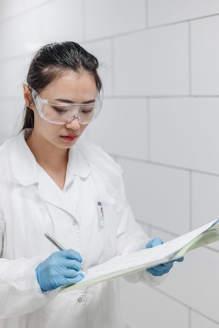 Asian female scientist wearing protective gear writes notes in a laboratory setting, focusing on research documentation.