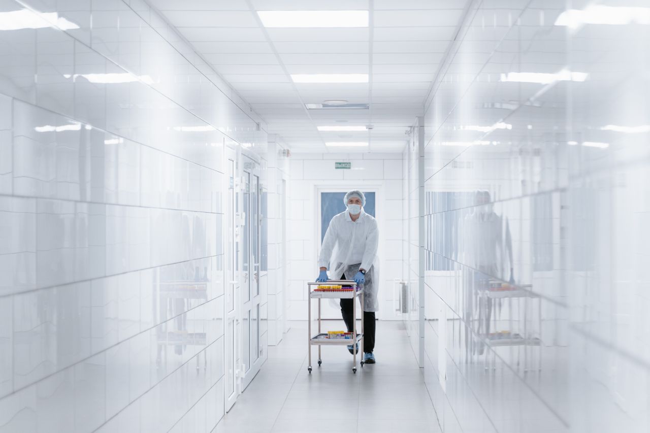 Scientist in protective gear moving samples on a cart through a bright laboratory hallway.